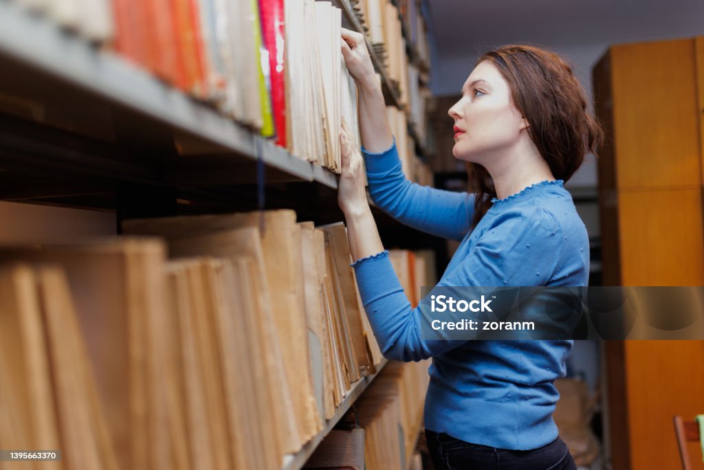 Female post-grad searching for study material, standing by the library shelves Nontraditional female student standing by the shelves in university library and looking for research material, adult student in continuing education Paper Stock Photo