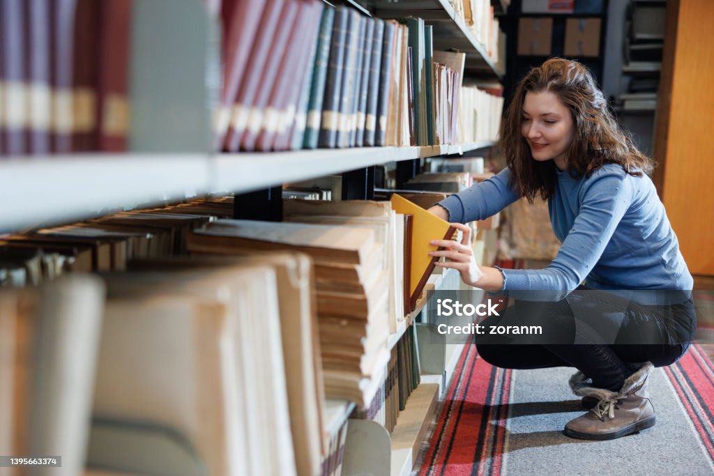 Female post-grad crouching by the shelves in university library, searching for material Nontraditional female student crouching by the lower shelves in university library, smiling and looking for research material, adult student in continuing education File Folder Stock Photo