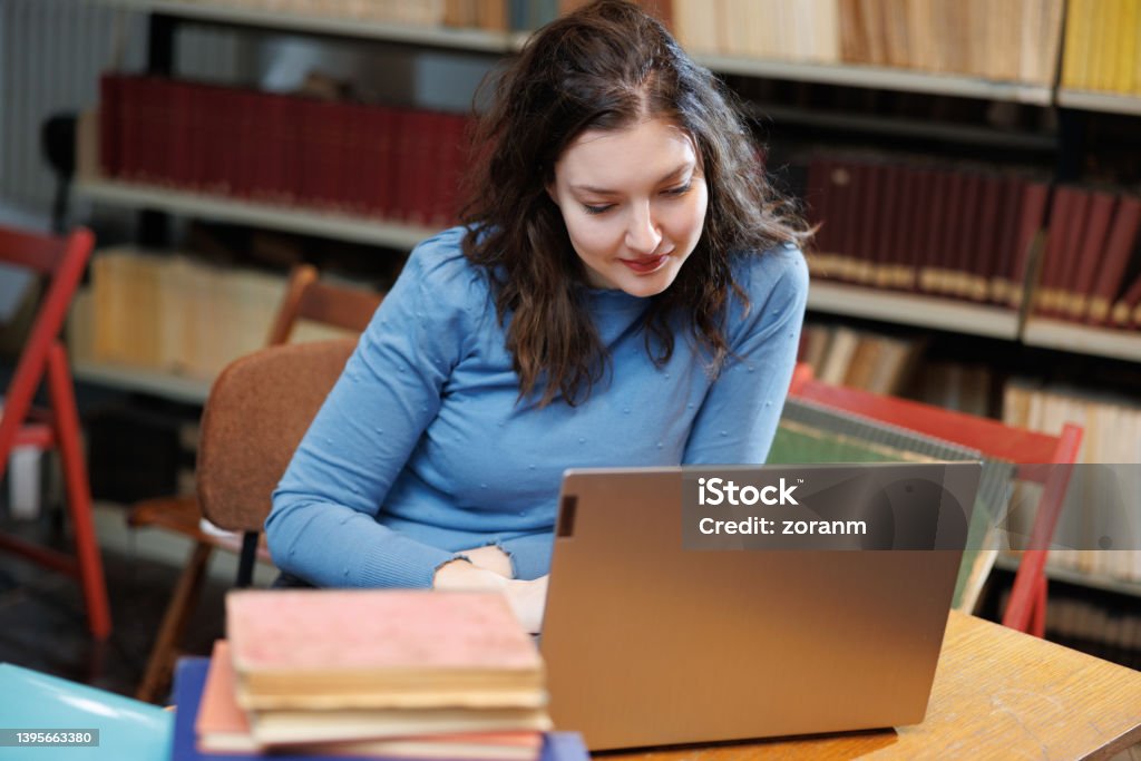 Beautiful post-grad using laptop at the desk in university library Female university student using laptop at the desk with a stack of selected books in university library 30-34 Years Stock Photo