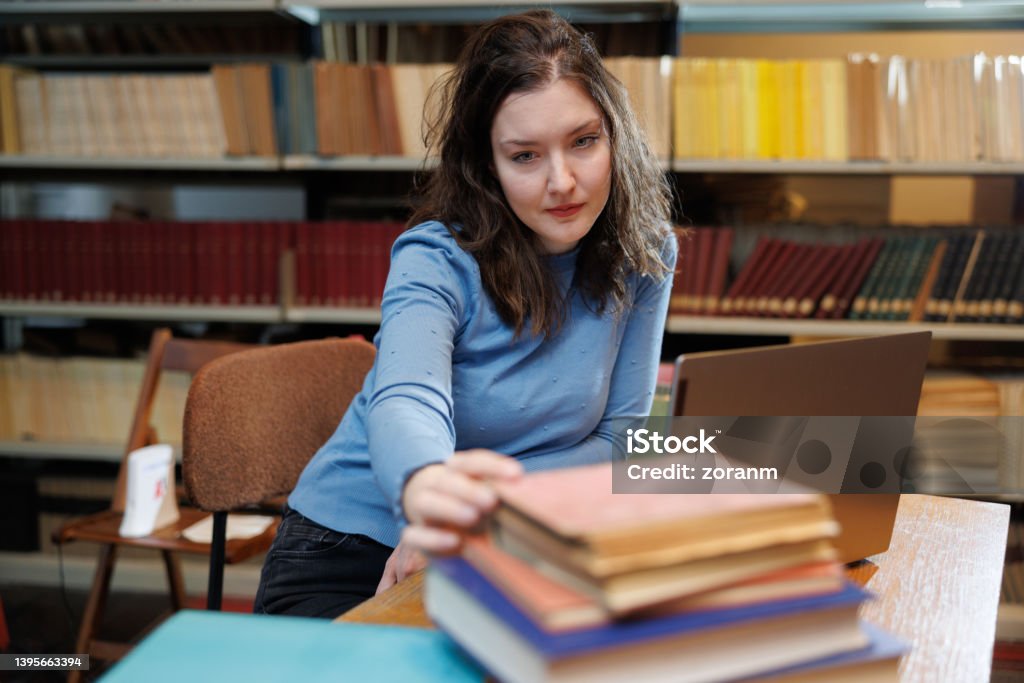 Female post-grad using laptop and picking up book from stack on the desk in library Beautiful university student using laptop in university library and picking up book from stack on the desk 30-34 Years Stock Photo