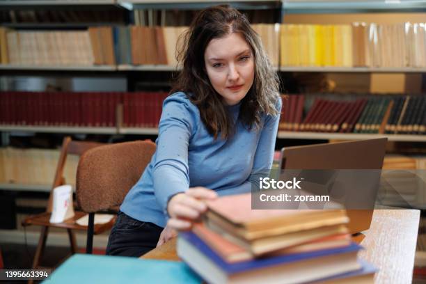 Female Postgrad Using Laptop And Picking Up Book From Stack On The Desk In Library Stock Photo - Download Image Now
