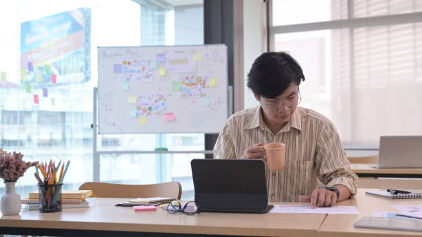 istock Handsome asian male manager holding coffee cup and working at modern office. 1395668088
