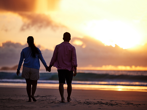 Back view of a romantic couple holding hands while walking towards the sunset at the beach. Only silhouettes are visible. The sky is orange. Romantic date on the beach in Santorini.