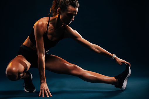 Athlete doing stretching exercises on black background. Female runner stretching leg muscles by touching his shoes while sitting in studio.