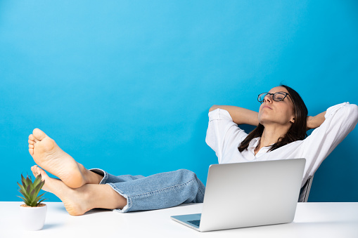 Pretty hispanic young woman relaxing feet on desk of office in front of laptop isolated on blue background.
