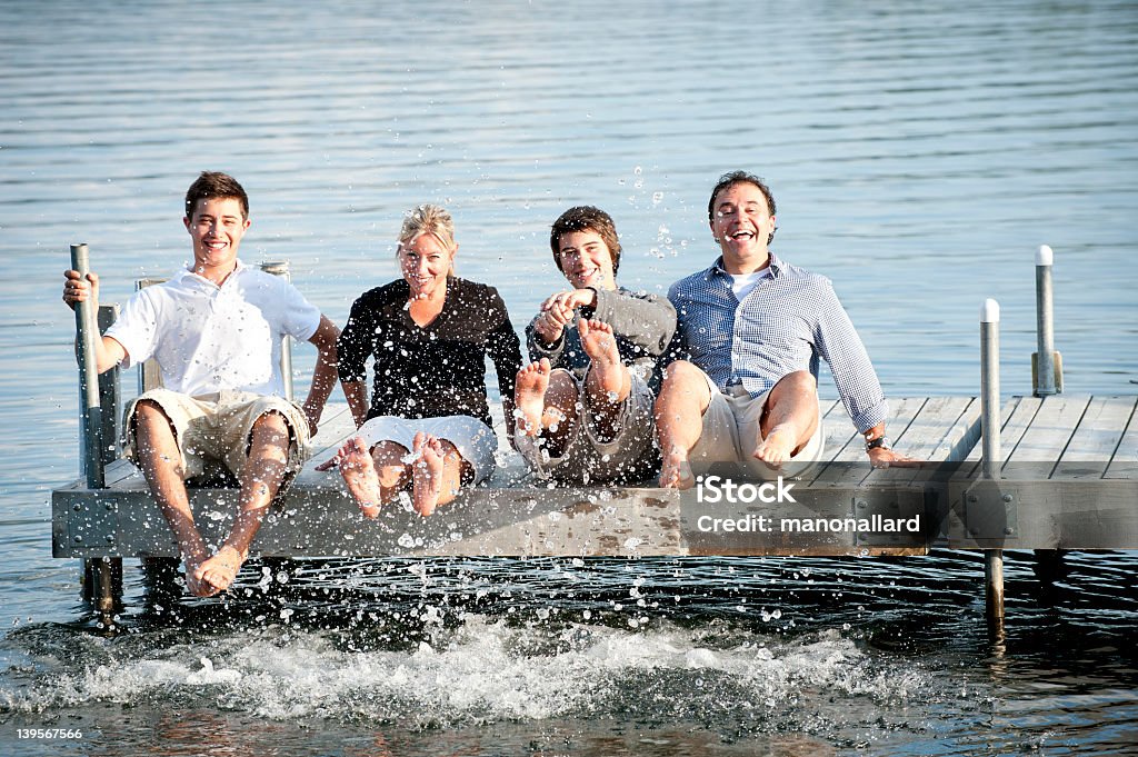 Happy family having fun playing in water with their feet Father Mother and sons sitting on a wooden pier on a lake have fun and playing in the water with their feet. 16-17 Years Stock Photo