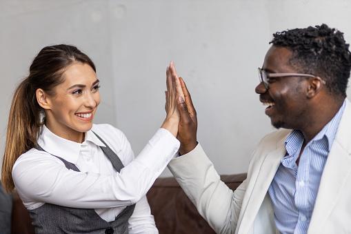 Beautiful young white woman and handsome black man celebrating after a business meeting. High-five between colleagues. Multiracial coworkers celebrating success at work.
