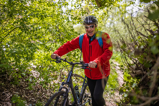 Active and excited senior man, pushing bicycle through forest, wearing cycling helmet