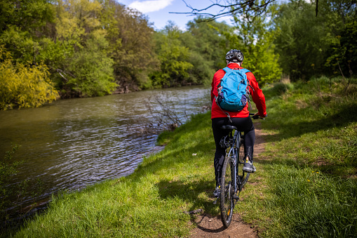 Rear view of active and healthy senior man, riding bicycle through forest near river, wearing cycling helmet