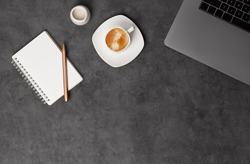 Workspace top view with laptop, notepad, pens and coffee on dark grey stone background. Flat lay with copy space. Business concept.