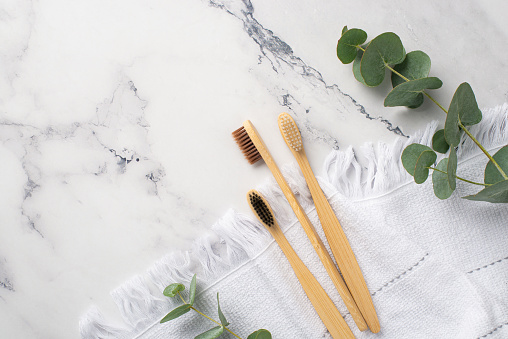 No plastic concept. Top view photo of three bamboo toothbrushes on white towel and eucalyptus on white marble background with copyspace