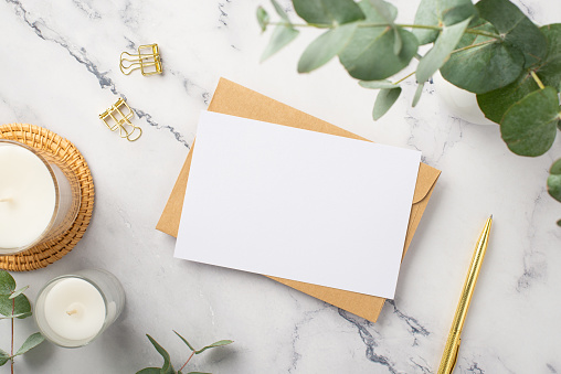 Business concept. Top view photo of candles on rattan serving mat paper sheet craft paper envelope gold pen binder clips and ceramic vase with eucalyptus on white marble background with copyspace