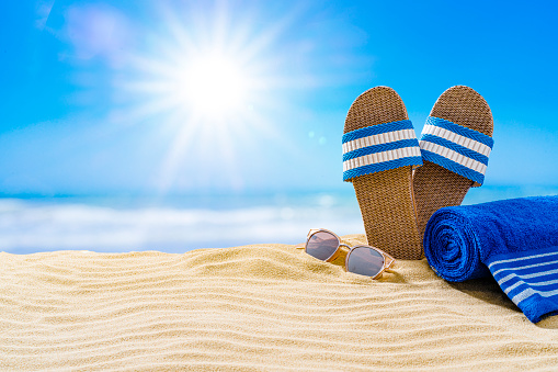 Summer accessories: sandals, beach towel and sunglasses on gold colored sand dune. The composition is at the right of an horizontal frame leaving useful copy space for text and/or logo at the left. High resolution 42Mp outdoors digital capture taken with SONY A7rII and Zeiss Batis 40mm F2.0 CF lens