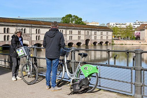 Strasbourg, France - April 2022: Two people sightseeing using bikes rented from the city's biycycle sharing system. In the background is the Barrage Vauban
