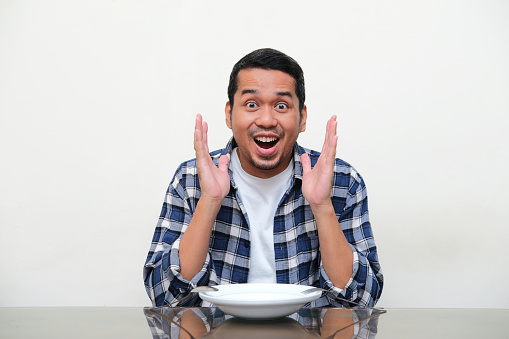 Adult Asian man sitting in front of empty plate and showing wow expression