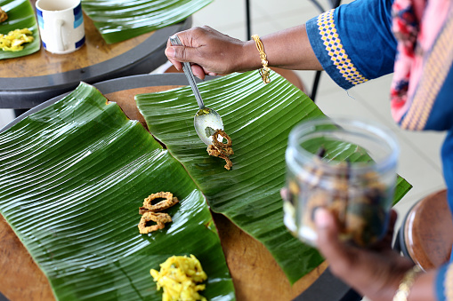 An Asian woman is putting side dishes on banana leaf for lunch preparation. Banana leaf is used as disposable plate for aromatic flavour.