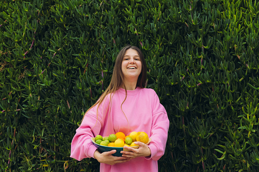 Portrait of a smiling female in a bright pink sweater staying by the green wall of plants holding a bowl of fresh colourful citrus fruits harvested from her garden