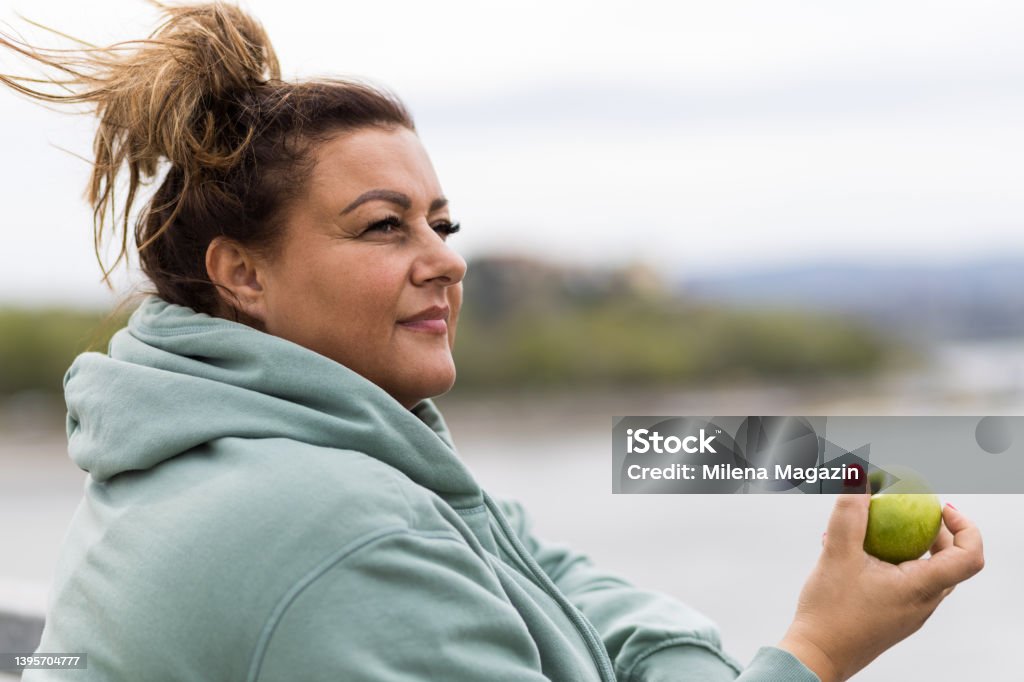 Portrait of an obese woman with an apple Overweight Hispanic woman leaning on a fence, holding a green apple after running cardio training. Obese woman exercising. Fat woman exercising. Body positivity. Plus size woman exercising. Weight loss journey. Obesity Stock Photo