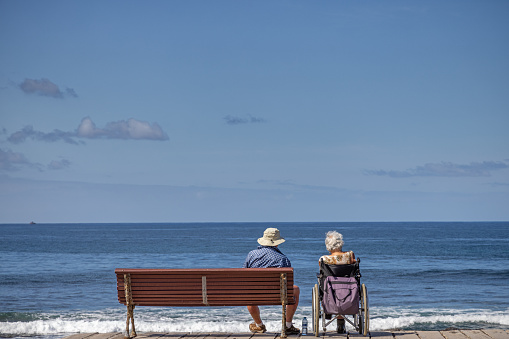 Man on a bench and his wife in a wheelchair is enjoying the view over the Atlantic Ocean at Playa de las Americas which is a popular tourist location on the south coast of the the Spanish Canary Island Tenerife.