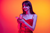 Portrait of young emotive girl, student using phone isolated over orange studio background in neon light. Concept of emotions, facial expression, art, beauty