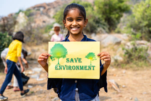 Happy smiling kid holding save environment sign board while other kids cleaning around at hill top - concept of support for climate change, sustainable lifestyle and volunteers.