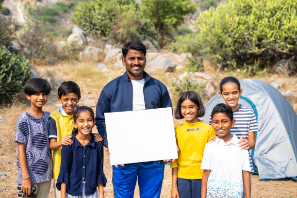 uśmiechnięta nauczycielka z nastoletnimi dzieciakami pokazująca białą pustą tablicę patrząc na kamerę na szczycie góry - koncepcja reklamy, trekkingu i promocji na obóz letni. - india mountain child people zdjęcia i obrazy z banku zdjęć