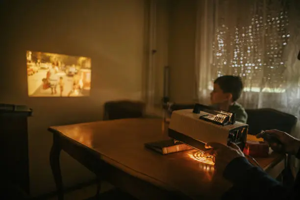 Grandfather and grandson sitting at the table and looking photos on vintage slide projector at elderly man's home.