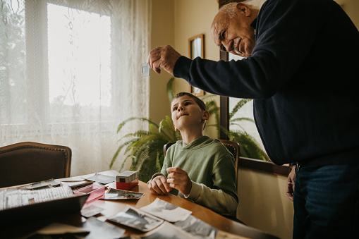 Grandfather and grandson sitting at the table and looking at photographic slides at elderly man's home.