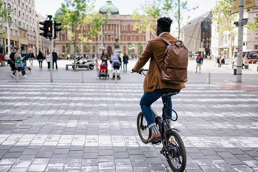 A behind view of a young entrepreneur going to work with his electrical bicycle, crossing the street on a pedestrian crossing in the city