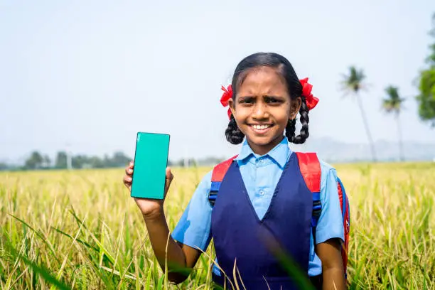 Happy smiling village girl kid showing green screen mobile phone while standing at paddy farm field - concept of advertisement, app promotion and education.