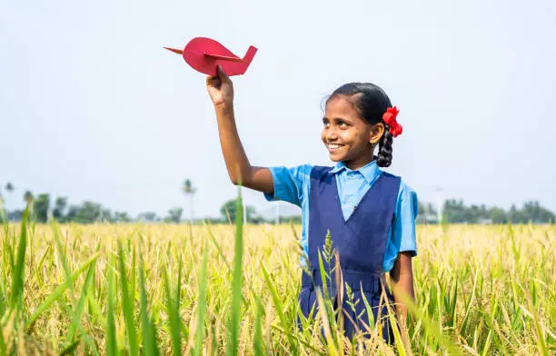 Photo of Happy village girl kid with school uniform playing using toy cardboard aeroplane at paddy field - concept of childhood dream, aspirations and freedom.