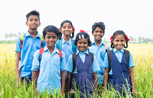 group of happy smiling village school kids in uniform standing at middle paddy field by looking camera - concept of friendship, education and togetherness.