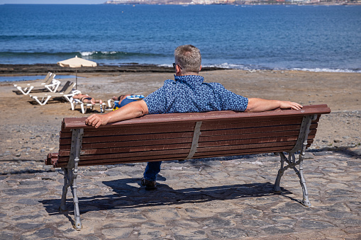 Man sitting on a bench and watching life on the beach at Playa de las Americas which is a popular tourist location on the south coast of the the Spanish Canary Island Tenerife.