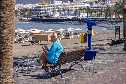 Woman having a break and checking her phone with a view to the Atlantic Ocean at Playa de las Americas which is a popular tourist location on the south coast of the the Spanish Canary Island Tenerife.