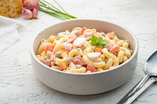 Homemade macaroni salad with elbow pasta, onion, carrot, tomato, green peas and mayonnaise dressing in a white bowl on a white wooden table