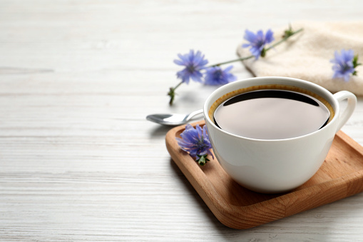 Cup of delicious chicory drink and flowers on white wooden table. Space for text