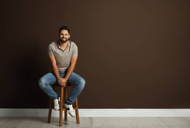 istock Handsome young man sitting on stool near brown wall. Space for text 1395742268