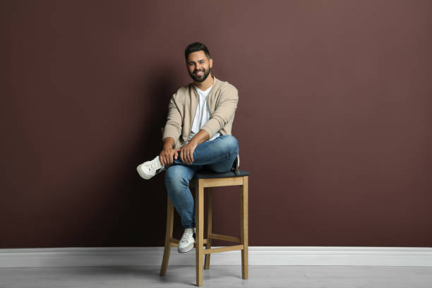 istock Handsome young man sitting on stool near brown wall 1395742269