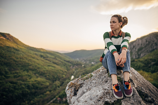 One woman, young female hiker sitting on a rock high on mountain in sunset.