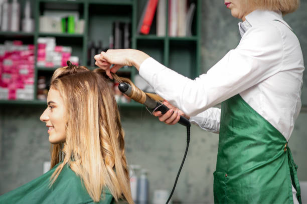 criando cachos com ferros enrolados. cabeleireiro faz um penteado para uma jovem com cabelos longos e ruivos em um salão de beleza. - mid teens hairstyle curly hair long hair - fotografias e filmes do acervo