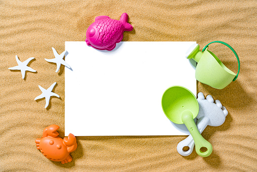 Kids summer backgrounds: overhead view of a blank card placed on golden sand background. Plastic beach toys are around the card. Copy space. High resolution 42Mp outdoors digital capture taken with SONY A7rII and Zeiss Batis 40mm F2.0 CF lens
