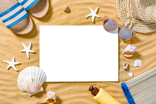 Summer backgrounds concept: overhead view of a blank card placed on golden sand background. Personal beach accessories like sandals, suntan lotion bottle, sunglasses sun hat and beach mat are around the card.  Starfish and sea shells complete the composition. Copy space. High resolution 42Mp outdoors digital capture taken with SONY A7rII and Zeiss Batis 40mm F2.0 CF lens