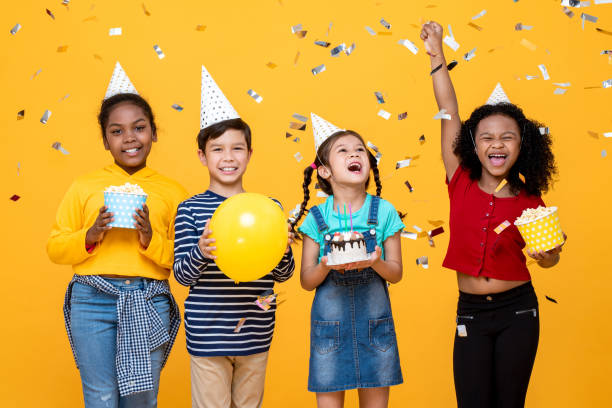 Multiethnic kids celebrating birthday party in studio yellow color background with confetti stock photo