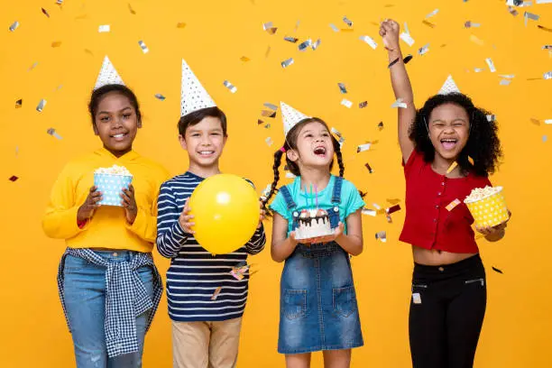 Photo of Multiethnic kids celebrating birthday party in studio yellow color background with confetti