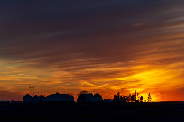 A setting sun reflects gold and yellow on clouds and silhouettes farm buildings on the horizon.