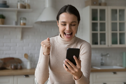Happy young woman look at phone screen clenched fist, scream with joy, rejoicing success, get great unbelievable news in message or sms sit in kitchen at home. Moment of win, luck, bet victory concept