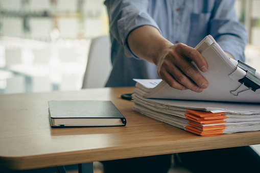 An employee sits at a table full of paper. stamp on the pile of unfinished documents A young account manager's secretary works in the office among piles of paperwork on the table.