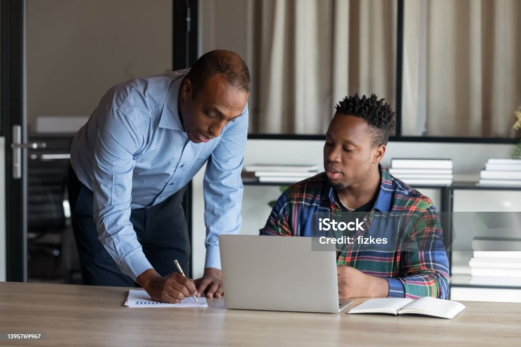 Serious African American adult mentor training intern at laptop Serious African American adult mentor training young millennial intern at laptop, explaining. Boss explaining work task to new employee at workplace. Coworkers discussing project issues in office Mentorship Stock Photo