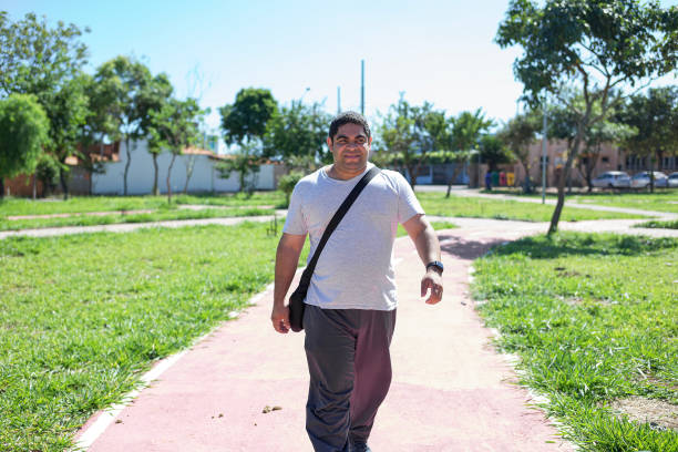 Portrait of a young man doing some exercises at a park, in a non urban city, surrounded by trees and a summer blue sky. He is walking on a lane and appreciating the nature around him.