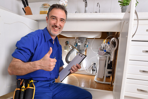 Plumbing technician checking water installation under the sink of a home kitchen with notepad and hand with ok gesture. Horizontal composition.